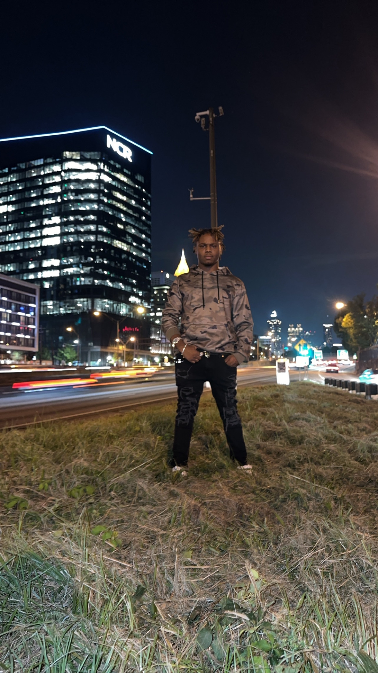 BABYSCAR in a camo hoodie at night beside a highway, city towers lit behind him