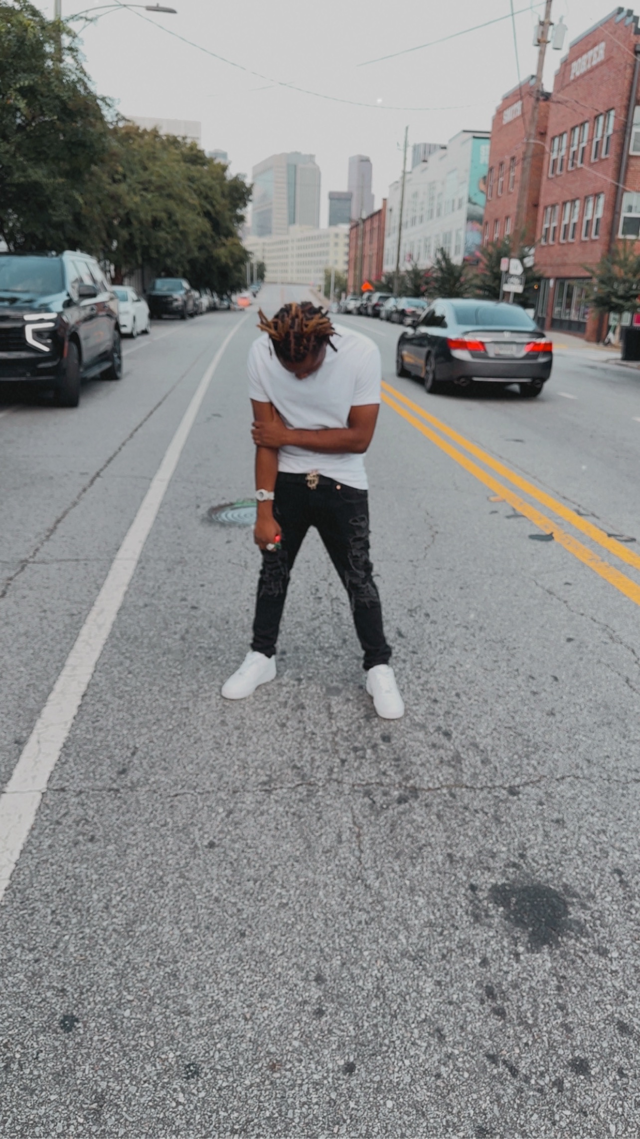 BABYSCAR standing in the middle of a city street in daylight, head bowed, arms crossed