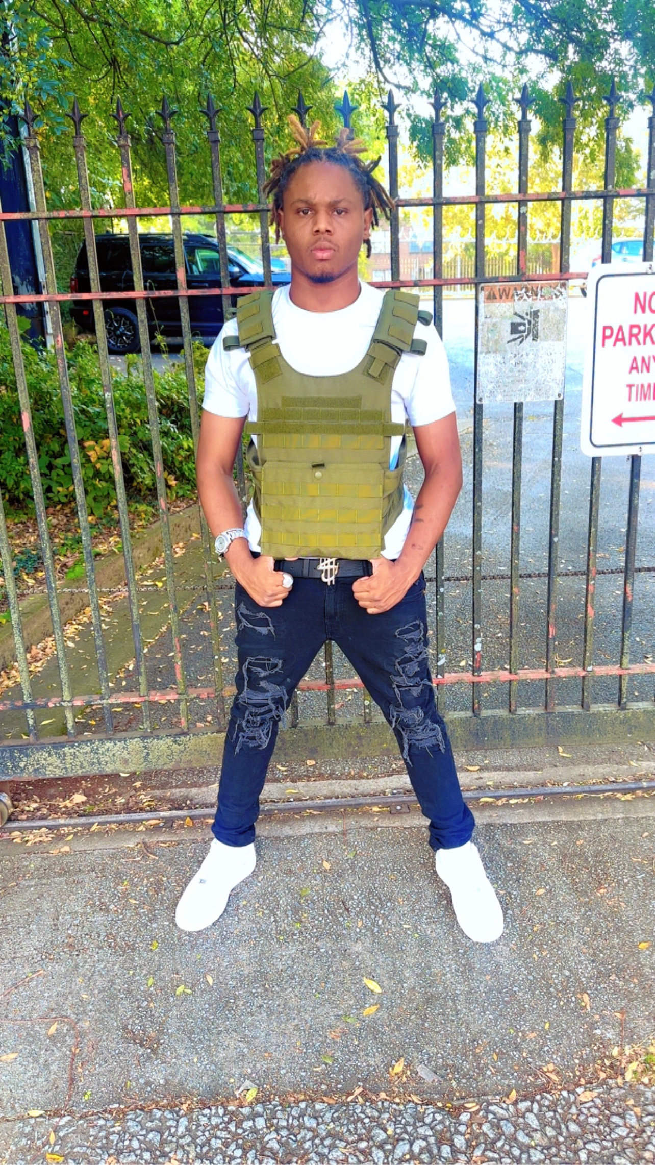BABYSCAR in a white tee and olive tactical vest, standing in front of an iron fence